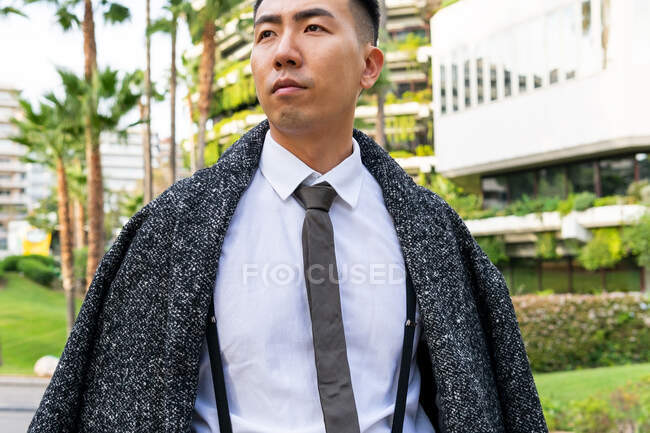 Gut gekleidete junge asiatische männliche Unternehmer mit Krawatte schauen weg, während sie auf der Straße gegen moderne Gebäude in der Stadt bummeln — Stockfoto