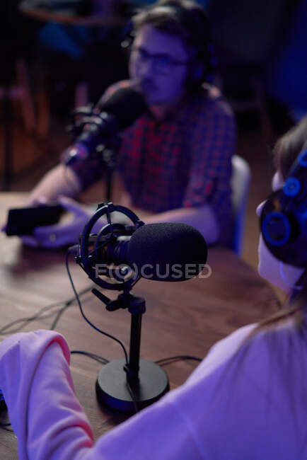 Concentrated young male and female colleagues in casual clothes and headphones sitting at table with microphones and communicating while recording podcast in modern studio — Stock Photo