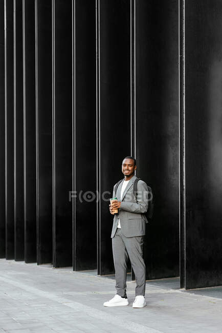 Позитивный афроамериканец с рюкзаком, стоящим с одноразовой чашкой кофе и смотрящим в сторону с зубастой улыбкой — стоковое фото