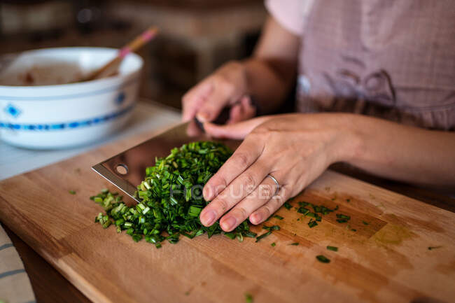 Von oben bis zur Unkenntlichkeit zerkleinert eine Frau frische grüne Kräuter auf einem Holzschneidebrett, während sie in der Küche das Abendessen zubereitet — Stockfoto
