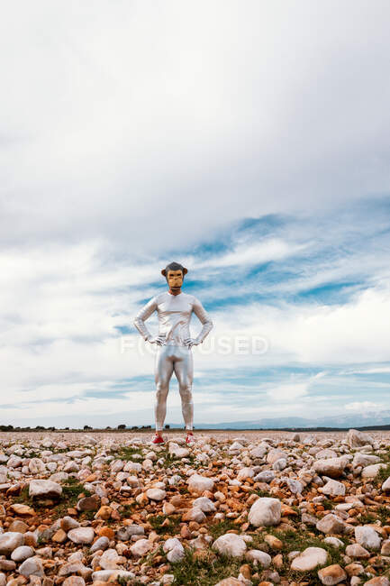 Male in monkey mask and silver latex outfit standing on stony field and looking at camera against cloudy blue sky — Stock Photo
