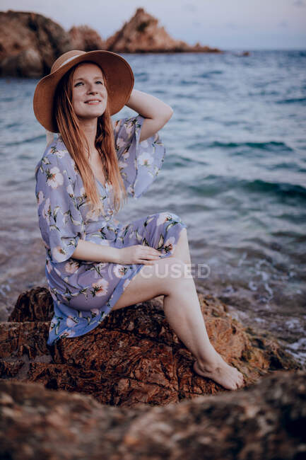 Charming young female in summer dress and hat sitting on rocky seashore while looking away in summer evening — Stock Photo