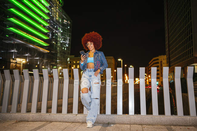 Ganzkörper positive Frau mit Afro-Frisur trägt trendige Outfit Textnachrichten auf dem Handy, während sie in der Nähe von Zäunen auf der Straße mit Gebäuden in der Abendzeit stehen — Stockfoto