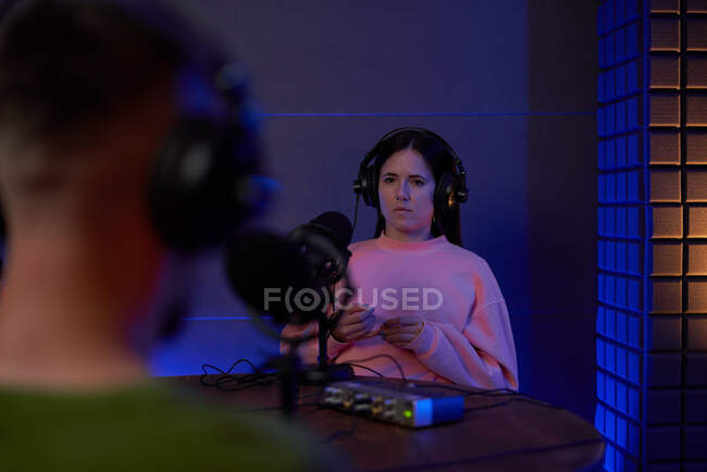 Joven presentadora de radio en ropa casual y auriculares sentada en la mesa con micrófono y comunicándose con su colega anónima durante la grabación de podcast en el estudio - foto de stock