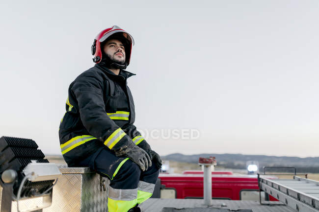 Hombre adulto pensativo que usa un sombrero protector y un traje protector mientras está sentado en la parte superior del camión de bomberos y mira hacia otro lado con el cielo despejado en el fondo - foto de stock
