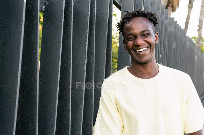 Alegre hombre afroamericano con camiseta casual de color amarillo claro y pelo corto parado en la valla y riendo mientras mira la cámara - foto de stock