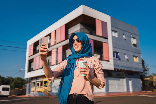 Femme arabe à la mode dans le hijab traditionnel debout avec du café pour aller dans la rue et la messagerie sur téléphone portable par une journée ensoleillée en ville — Photo de stock