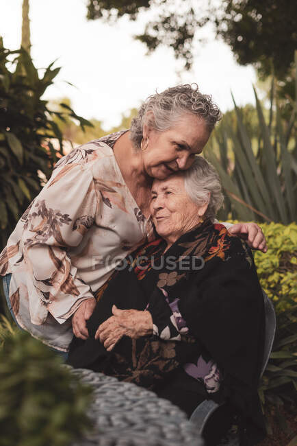 Feliz anciana abrazando y besando a la hermana en la cabeza y luego sonriendo mientras pasan tiempo en el jardín juntos - foto de stock