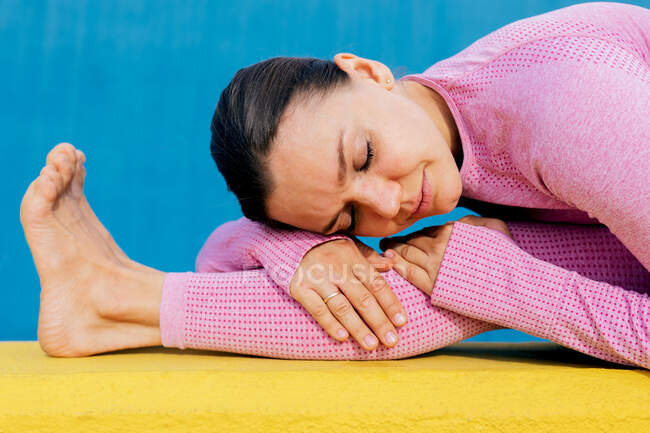 Молодая женщина в розовой спортивной одежде сидит с закрытыми глазами и растягивает ноги на желтом коврике — стоковое фото