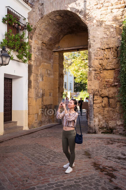 Corps complet de voyageuse asiatique prenant des photos sur son téléphone portable alors qu'elle se tenait debout sur une passerelle pavée près de bâtiments âgés et d'une arche en pierre en ville — Photo de stock