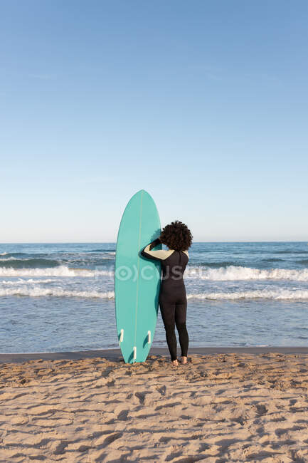 Visão traseira de surfista feminina irreconhecível em roupa de mergulho com prancha de surf de pé olhando para longe na praia lavada pelo mar ondulando — Fotografia de Stock