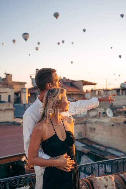 Loving man embracing woman from behind and pointing away on roof terrace with hot air balloons in evening sky in Cappadocia Turkey — Stock Photo