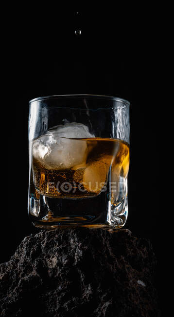 Whiskey-Tropfen fallen auf Eiswürfel, die in Kristallglas auf rauer Oberfläche vor schwarzem Hintergrund serviert werden — Stockfoto