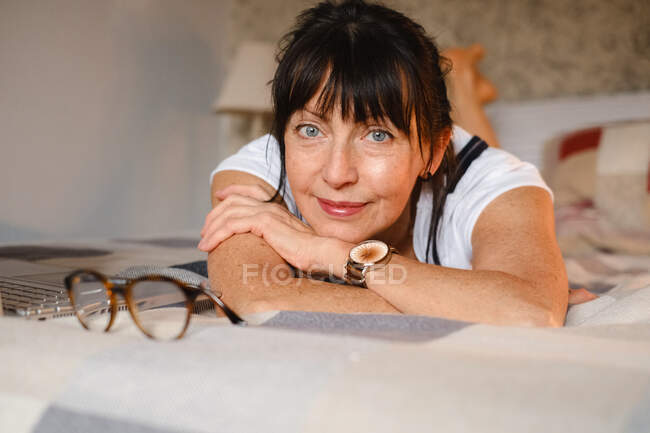 Позитивна жінка середнього віку спирається на руку, лежачи на ліжку з відкритою блокнотом та окулярами і дивлячись на камеру — стокове фото