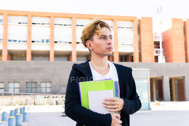 Calm male student with workbooks looking away with thoughtful face while standing near modern university building on street during studies — Stock Photo
