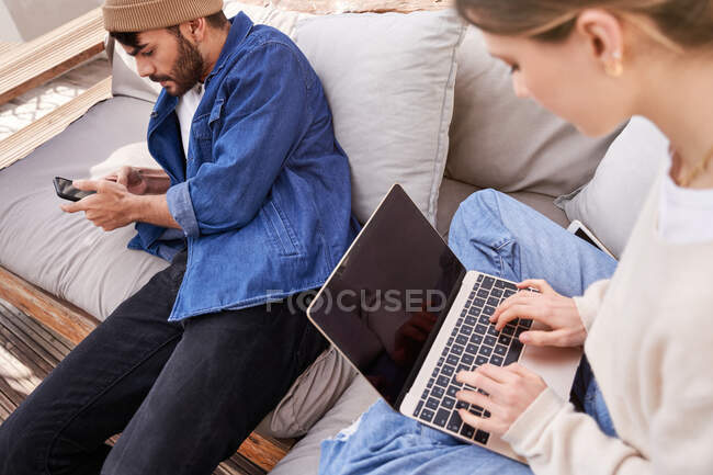 Hohe Winkel der Ernte junge Frau sitzt mit gekreuzten Beinen und mit Netbook in der Nähe männlichen Mitbewohner scrollen Handy — Stockfoto