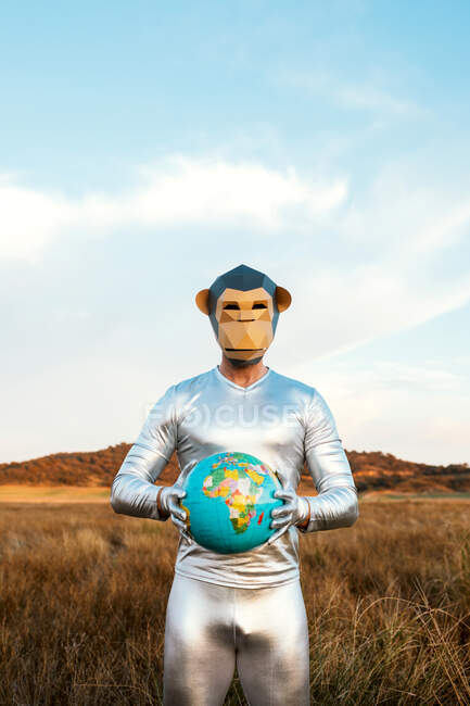 Homem anônimo em terno de látex de prata com máscara de macaco geométrico olhando para a câmera e segurando globo na natureza — Fotografia de Stock
