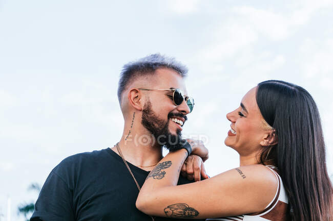Mujer feliz con el pelo negro en ropa casual abrazando novio alegre en gafas de sol mientras se ríe en la calle durante el día - foto de stock