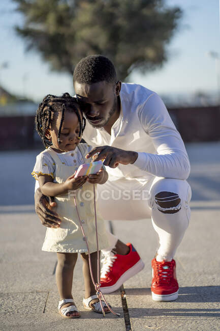 Афроамериканський батько у сорочці, присідає з маленькою дівчинкою з зачіскою на вулиці, а в сонячний день дивиться на смартфон. — стокове фото