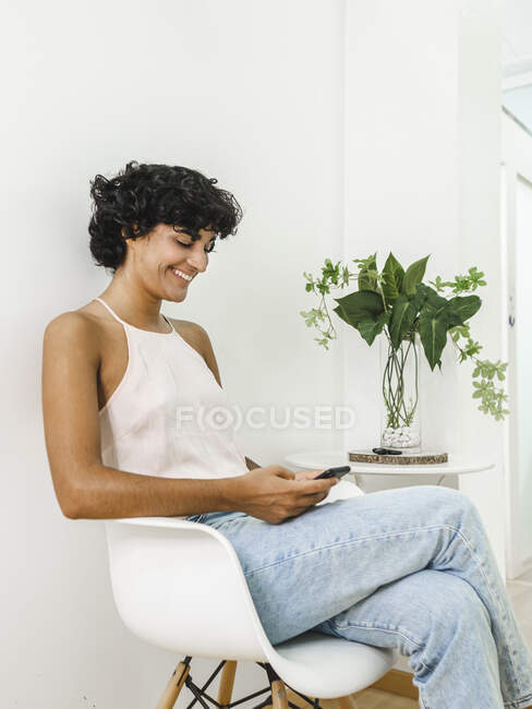 Веселая латиноамериканка сидит в кресле и просматривает мобильный телефон в светлой комнате рядом с растением на столе — стоковое фото