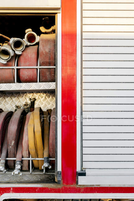 Section with various types of twisted fire hoses and different types of adaptors located on two shelves in fire truck and another section with the fire truck shutter down in bright sunlight at daytime — Stock Photo