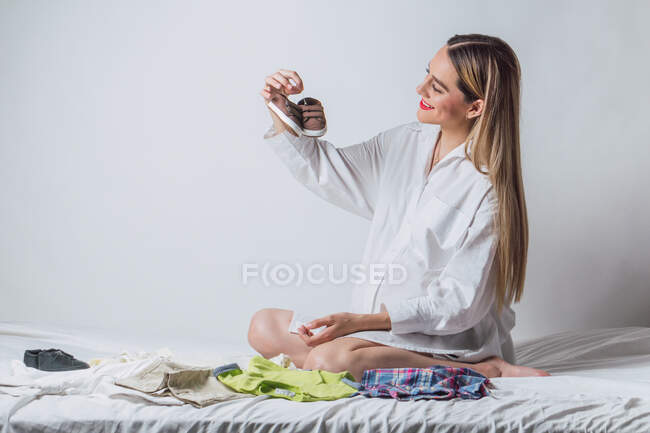 Mujer joven positiva con camisa blanca sentada en la cama y preparando ropa de bebé para el parto mientras está sentada en la cama - foto de stock