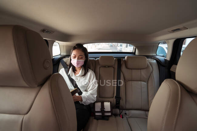 Женщина в формальной одежде в защитной маске записывается в блокнот во время езды на пассажирском заднем сидении в удобном такси — стоковое фото