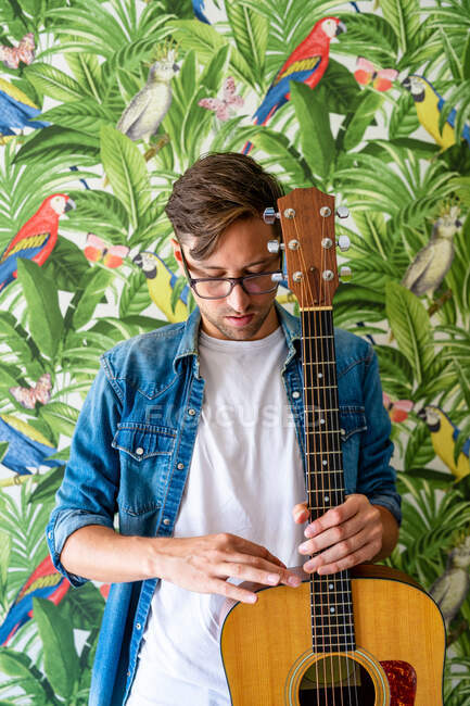 Concentrated male guitarist while standing against wall with vivid paintings of green plants and parrots — Stock Photo