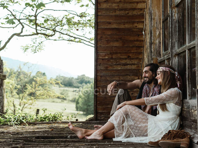 Vista lateral de um par de hippies sentados no piso do alpendre com os pés descalços olhando para longe — Fotografia de Stock