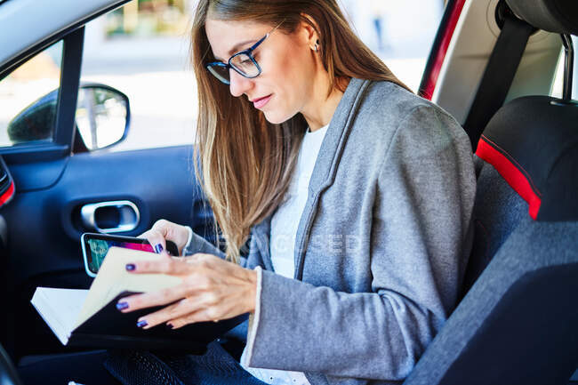 Empresaria ocupada sentada en el asiento del pasajero en el coche y tomando notas en el bloc de notas mientras usa el teléfono inteligente - foto de stock