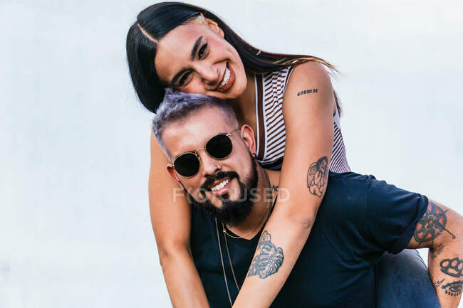 Delighted female with dark hair in casual clothes embracing smiling boyfriend with tattoos in sunglasses and looking at camera against light background — Stock Photo