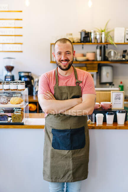 Позитивный мужчина-бариста в фартуке смотрит на камеру со скрещенными руками, стоя возле прилавка с бумажными чашками и десертами в кофейне — стоковое фото