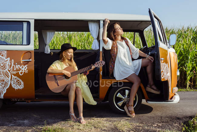 Due belle ragazze vestite in abiti estivi divertirsi fuori da un furgone e suonare la chitarra — Foto stock
