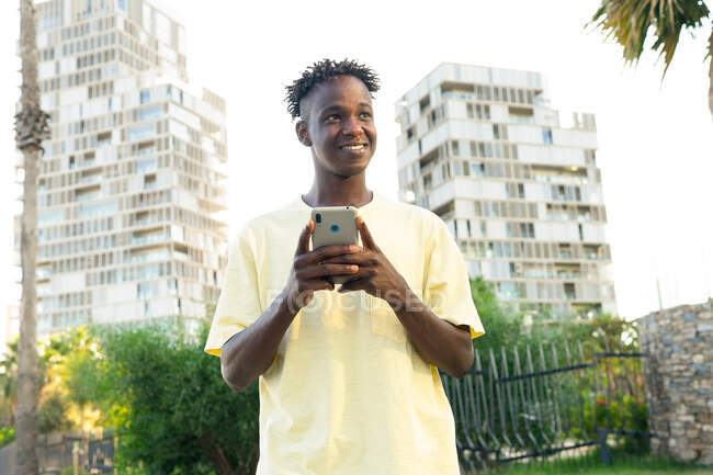 Un homme afro-américain souriant portant un t-shirt décontracté envoyant un message texte sur son téléphone portable et regardant loin en ville avec des paumes — Photo de stock