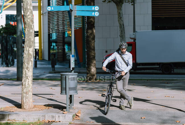 Longitud completa del trabajador masculino barbudo en casco que cruza la carretera de asfalto con bicicleta en camino al trabajo - foto de stock