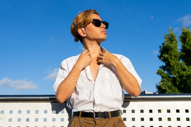 Seriöser junger Mann in weißem Hemd und moderner Sonnenbrille blickt in die Ferne, während er auf der sonnigen Straße mit dem Gebäude in der Stadt steht — Stockfoto