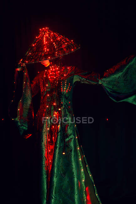 Неузнаваемая загадочная женщина в креативном традиционном наряде и вьетнамской головной убор с красной подсветкой, стоящая в темной студии на черном фоне во время выступления — стоковое фото