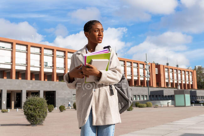 Umtriebige afroamerikanische Studentin mit einem Haufen Schulbücher schaut weg, während sie auf der Straße in der Nähe des Universitätsgebäudes steht — Stockfoto