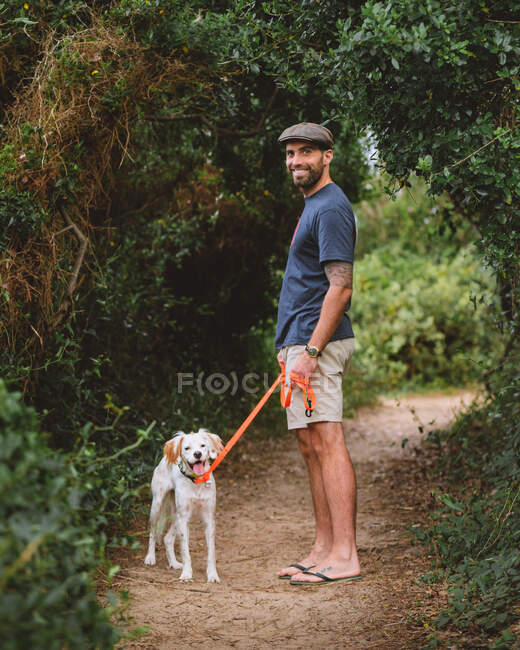 Full body of cheerful bearded male owner with cute dog on leash looking at camera while standing on path near tall green plants in nature — Stock Photo