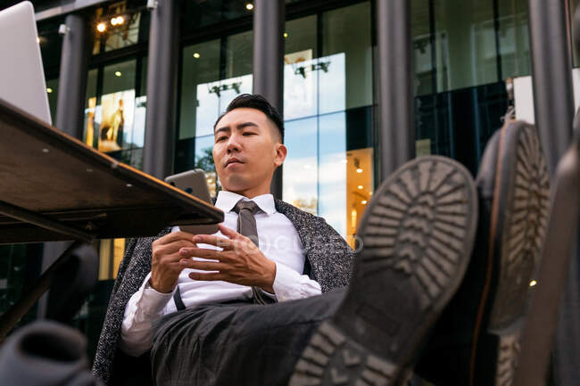 Atento joven empresario étnico sentado con las piernas cruzadas mientras navega por Internet en el teléfono celular en la mesa de la cafetería en la ciudad - foto de stock