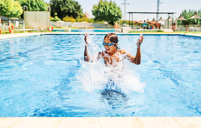 Niño feliz con el pelo mojado en gafas de natación saltando en la piscina mientras mira hacia otro lado y sonríe en el soleado día de verano - foto de stock