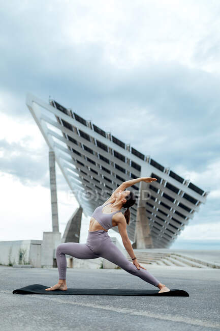 Corpo completo di donna sportiva in activewear praticare Viparita Virabhadrasana postura durante l'allenamento su strada vicino al pannello solare contro cielo nuvoloso — Foto stock