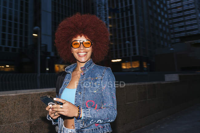 Attraktive Frau mit Afro-Frisur und trendigem Outfit, SMS auf dem Handy, während sie abends auf der Straße mit modernen Gebäuden steht — Stockfoto