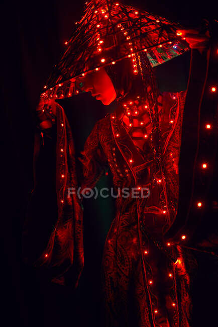 Enigmatic female in creative traditional outfit and Vietnamese headwear with red illumination standing in dark studio on black background during performance — Stock Photo