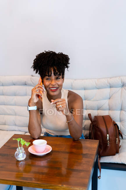 Entzückte junge Afroamerikanerin mit dunklem lockigem Haar in stilvoller Kleidung lächelt, während sie im Café am Tisch mit einer Tasse Kaffee telefoniert — Stockfoto