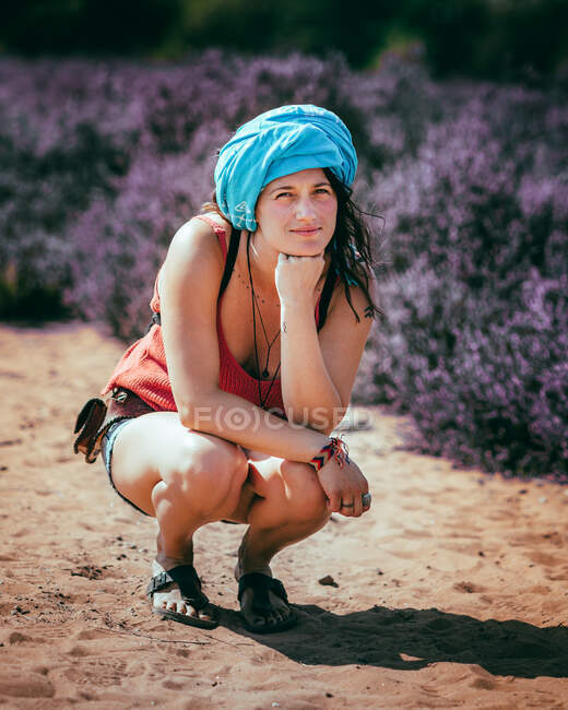 Mujer joven sonriente con cabello castaño en ropa casual y pañuelo en la cabeza agachándose y apoyándose en el campo de lavanda en el campo en un día soleado - foto de stock