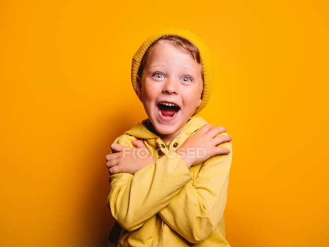 Joyeux petit garçon en imperméable à la mode et bonnet chapeau debout avec les bras croisés et regardant la caméra sur fond jaune en studio — Photo de stock