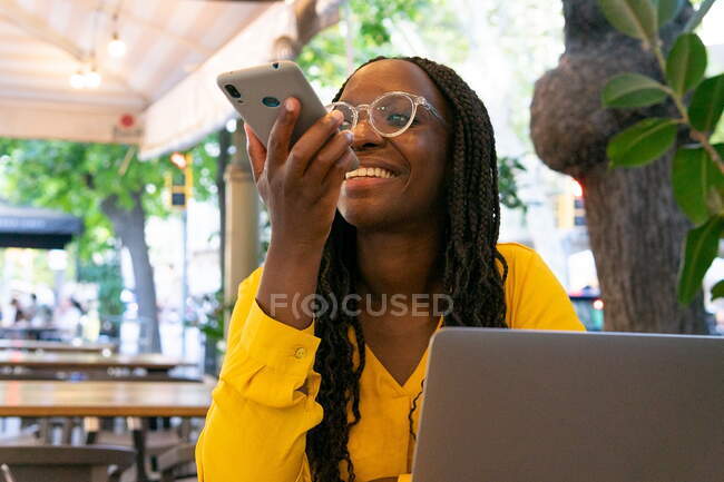 Positive African American female with black hair in sunglasses recording voice message on modern smartphone while sitting in cafe with plants — Stock Photo