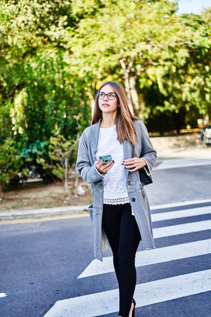 Серьёзная женщина в формальной одежде и пальто с сумочкой, пересекающей асфальтированную дорогу, при помощи мобильного телефона и отводя взгляд — стоковое фото