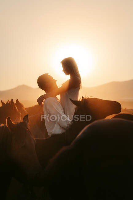 Vista lateral de la mujer feliz admirando la puesta de sol sobre las montañas mientras es criado por el hombre amoroso entre caballos tranquilos en el campo de Turquía - foto de stock